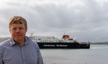 Tim stands near the water with a CalMac ferry in the background